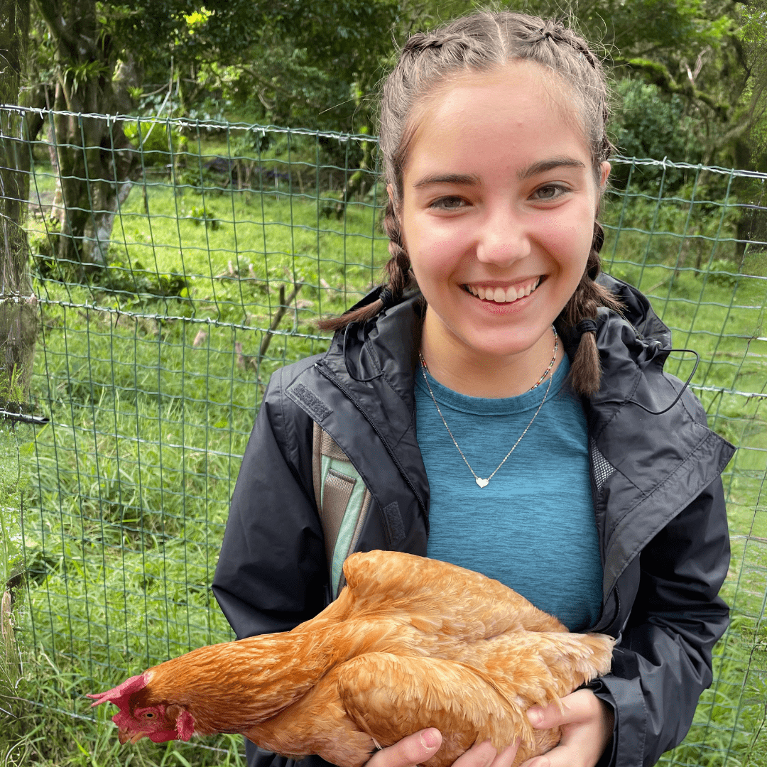 Natlie is standing in a grassy paddock, holding a hen and smiling at the camera. She is wearing a blue shirt, black open jacket, and has her hair in two french braids.